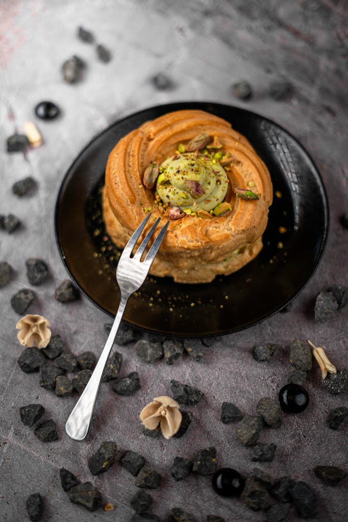 A beautifully presented pistachio cake topped with nuts and a fork, styled on a textured surface.