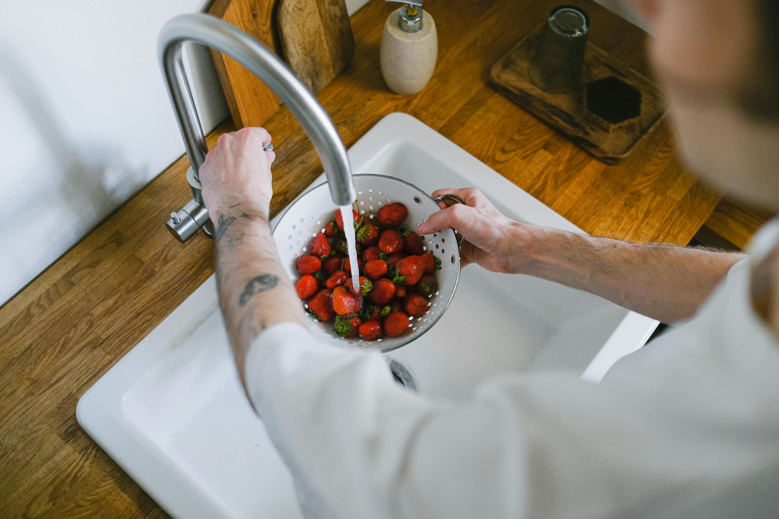 A person washes fresh strawberries in a kitchen sink using a strainer. Overhead angle captures the process.