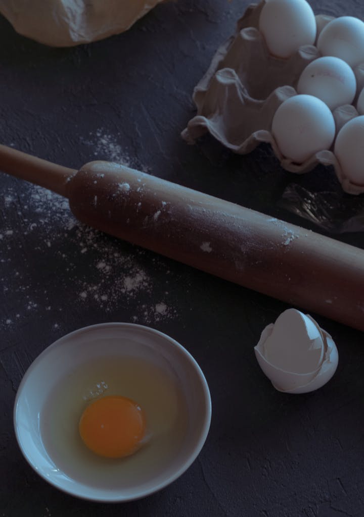 High angle view of baking ingredients like eggs and rolling pin on a dark kitchen table.